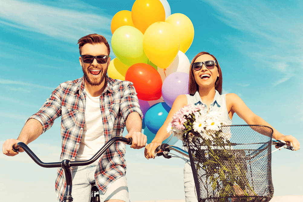 Una pareja maneja bicicleta felices con globos  de colores detrás de ellos